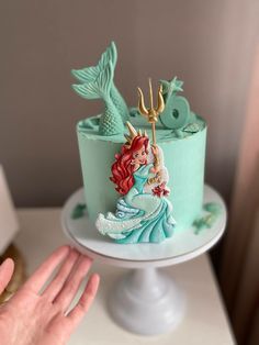 there is a little mermaid cake on top of the table with someone's hand next to it