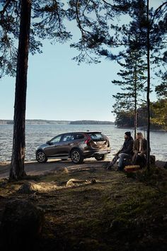 two people sitting on the ground next to a parked car near some trees and water