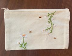 a white pouch with daisies and bees embroidered on the side, sitting on a wooden surface