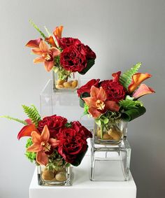 three clear vases with red flowers and greenery in them on a white table