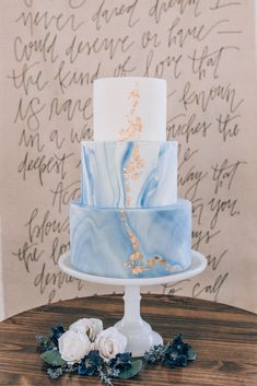 a three tiered blue and white cake on a wooden table next to some flowers