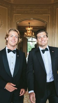 two men in tuxedos standing next to each other near a chandelier