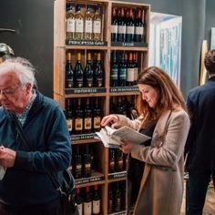 two people standing in front of a wine rack with bottles on it and one person reading a paper