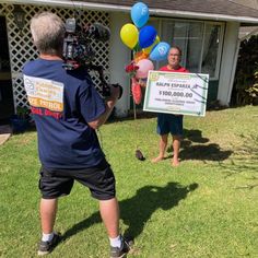 a man standing in front of a house holding a sign and balloons while another man is filming him