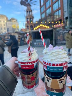 two people holding up cups with whipped cream and chocolate milkshakes in front of a building