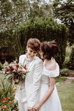 two women in white dresses standing next to each other and one is holding a bouquet