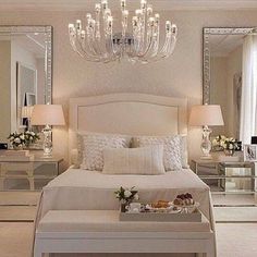 a white bedroom with chandelier, mirror and tray of food on the bed