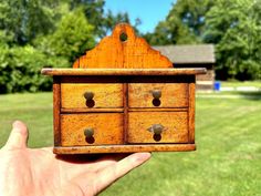 a hand holding a small wooden drawer in front of a grassy field with trees behind it