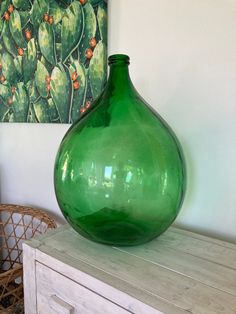 a large green vase sitting on top of a wooden table next to a painting and wicker chair