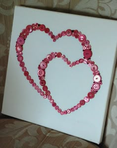 a heart made out of buttons sitting on top of a white canvas with pink trim