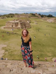 a woman standing on top of a stone wall next to a lush green field with ruins in the background