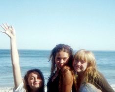 three girls standing on the beach with their arms in the air and one girl holding her hand up