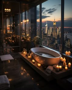 a bathtub with candles on the floor in front of a large window overlooking a city at night