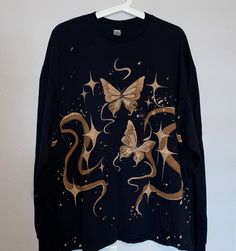 a black shirt with gold butterflies and stars on the front is hanging from a white hanger