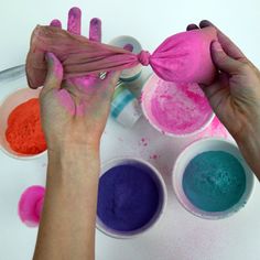 two hands are holding pink and blue powder in front of white bowls with different colors