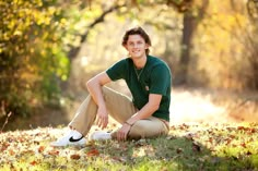 a young man is sitting on the ground in front of some trees and smiling at the camera