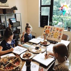 three children are sitting at a table with books and papers in front of them, while one child is drawing