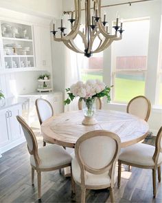 a dining room table with white chairs and plates on it, in front of a chandelier