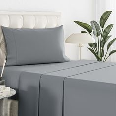 a bed with grey sheets and pillows next to a plant