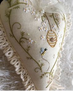 a white heart shaped pillow with flowers and bees on it's side, sitting on a furry surface