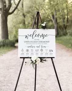 a welcome sign with flowers and greenery hanging from it's easel in the middle of a dirt road