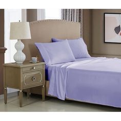 a bed with purple sheets and pillows in a room next to a lamp on a nightstand