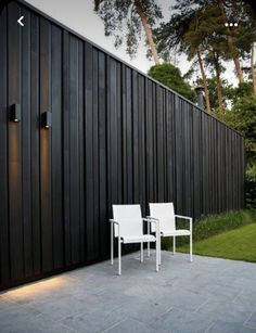 two white chairs sitting on top of a patio next to a black wall and grass