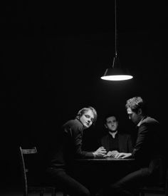 three men sitting at a table in the dark, one is looking at another man