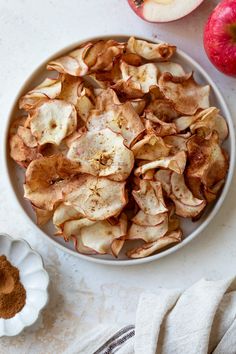 an apple chips in a white bowl next to some apples and cinnamon on a table