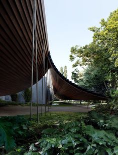 a curved wooden structure in the middle of some plants and trees, next to a building