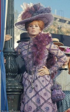 a woman in a purple dress and hat standing next to a lamp post with people looking on