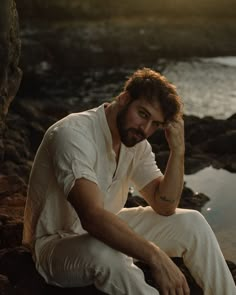 a man sitting on top of a rock next to the ocean with his hand near his face