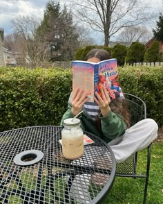 a person sitting at a table with a book in front of her face and holding a drink