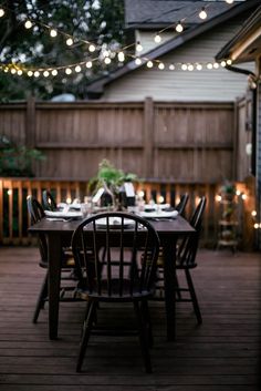 an outdoor dining area with lights strung over the table and wooden flooring, along with black chairs