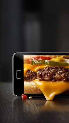 a hamburger with cheese and peppers on it being held up by a cell phone that is sitting on a table