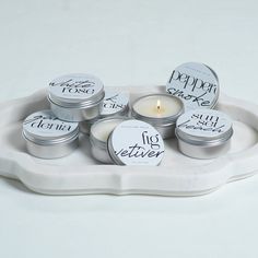 six small tins with labels on them sitting on a white tray next to a candle