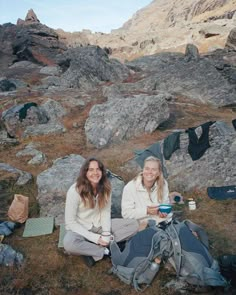 two women sitting on the ground next to some rocks and camping gear with mountains in the background