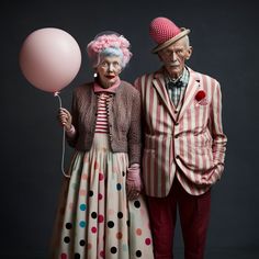 an old man and woman dressed up as clowns with balloons in their hands, standing next to each other