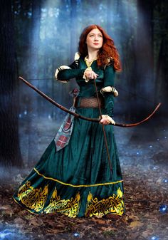 a woman dressed in green holding a bow and arrow while standing in the woods with her hands behind her back