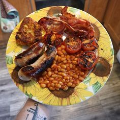 a plate with sausages, beans, tomatoes and other foods on it is being held up by someone's arm