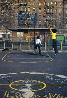 two young boys playing basketball on an outdoor court with graffiti written on the ground and buildings in the background