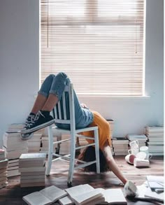 a woman laying on top of a wooden chair next to stacks of books in front of a window