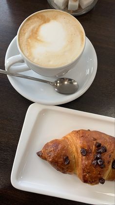 a croissant on a plate next to a cup of cappuccino