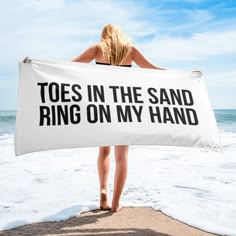 a woman standing on the beach holding a towel that says toes in the sand ring on my hand