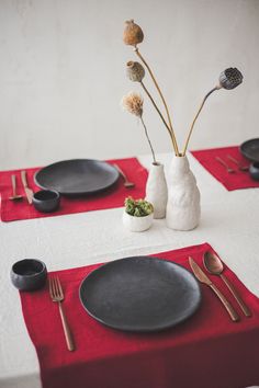 the table is set with black plates, silverware and flowers in vases on red placemats