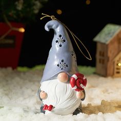 a gnome figurine sitting in the snow next to a house and christmas tree