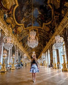 a woman is walking in an ornate hall with chandeliers and paintings on the walls