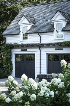 a white house with two black garage doors and some flowers in front of the building