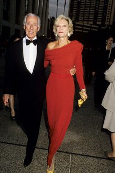 an older man and woman dressed in formal wear walking down the street with their arms around each other