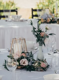 an arrangement of flowers and greenery on a white table cloth with candles, plates and wine glasses